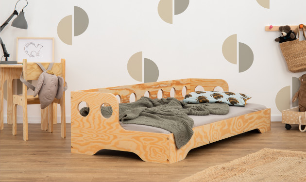 Drewniane łóżko z barierką o okrągłych wycięciach na długim boku wykonane ze sklejki sosnowej w kolorze lakierowanego drewna